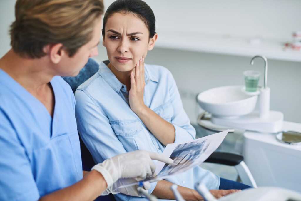 Woman in dental chair speaking with dentist holding hand to jaw in pain